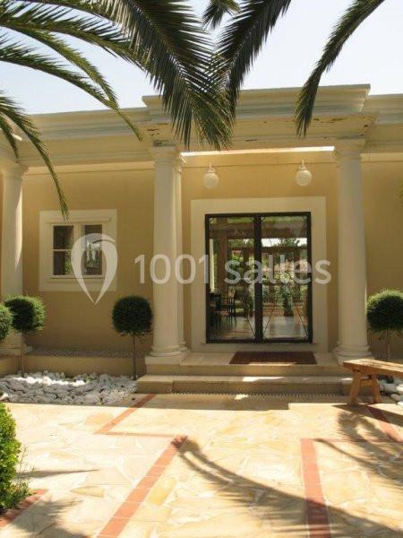 Entrée d'une maison beige avec colonnes blanches, porte vitrée, plantes taillées et allée pavée sous des palmiers.