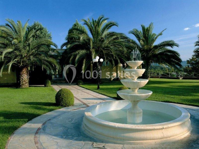 Fontaine à plusieurs niveaux entourée de pelouses, palmiers et arbustes dans un jardin ensoleillé.