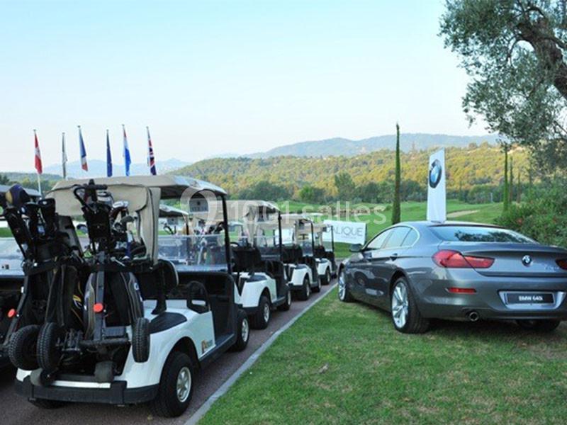 Voitures de golf alignées près d'une berline grise sur un terrain verdoyant avec des drapeaux en arrière-plan.
