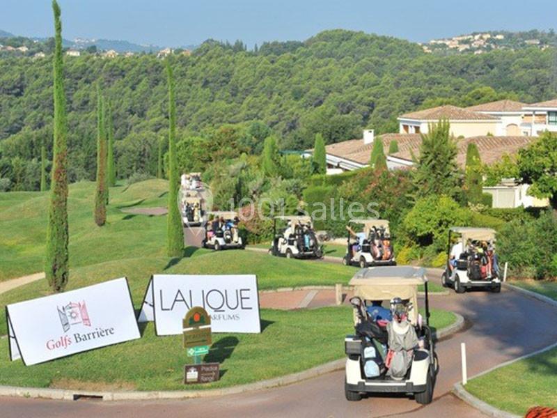 Des voiturettes de golf circulent sur un chemin entouré de pelouses et de bâtiments, avec des panneaux promotionnels…