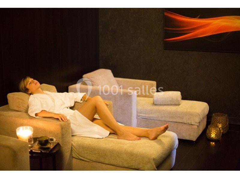 Femme en peignoir allongée sur une chaise longue dans un espace détente avec lumière tamisée et bougie allumée.