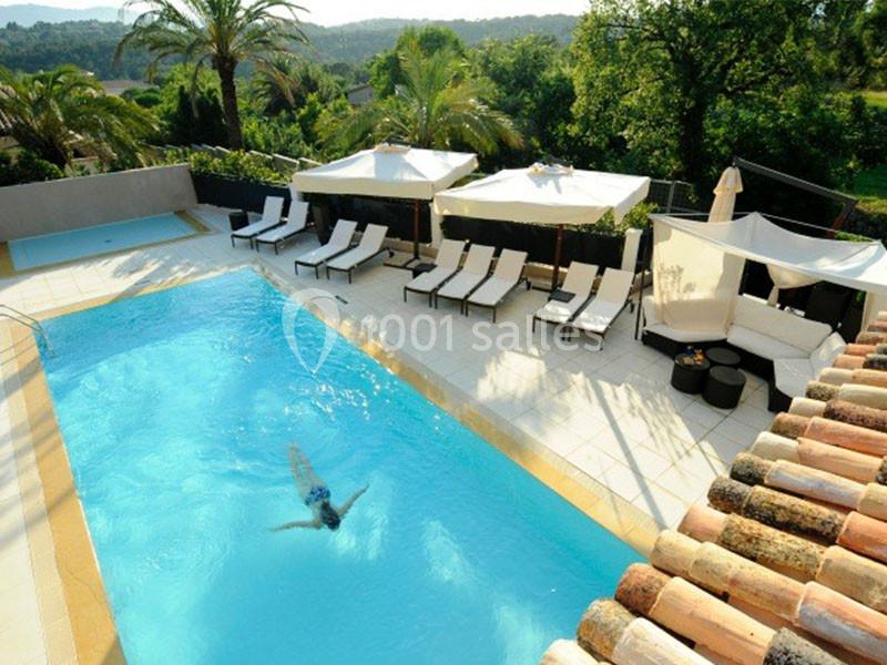 Piscine extérieure entourée de transats, parasols et végétation, avec une personne nageant au centre.