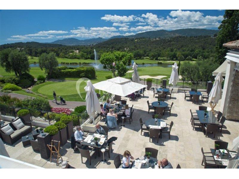 Terrasse d'un restaurant avec des tables occupées, vue sur un golf verdoyant et des collines sous un ciel dégagé.