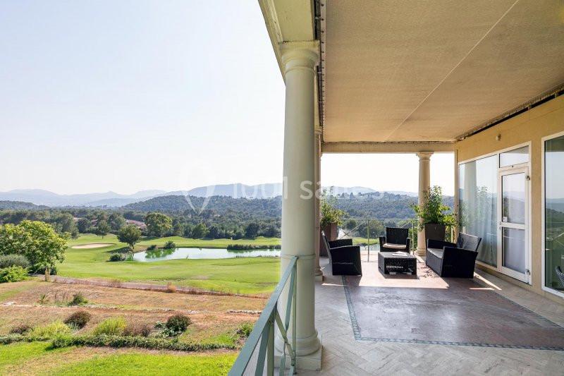 Vue depuis une terrasse couverte avec mobilier d'extérieur, donnant sur un terrain de golf entouré de collines verdoyantes.