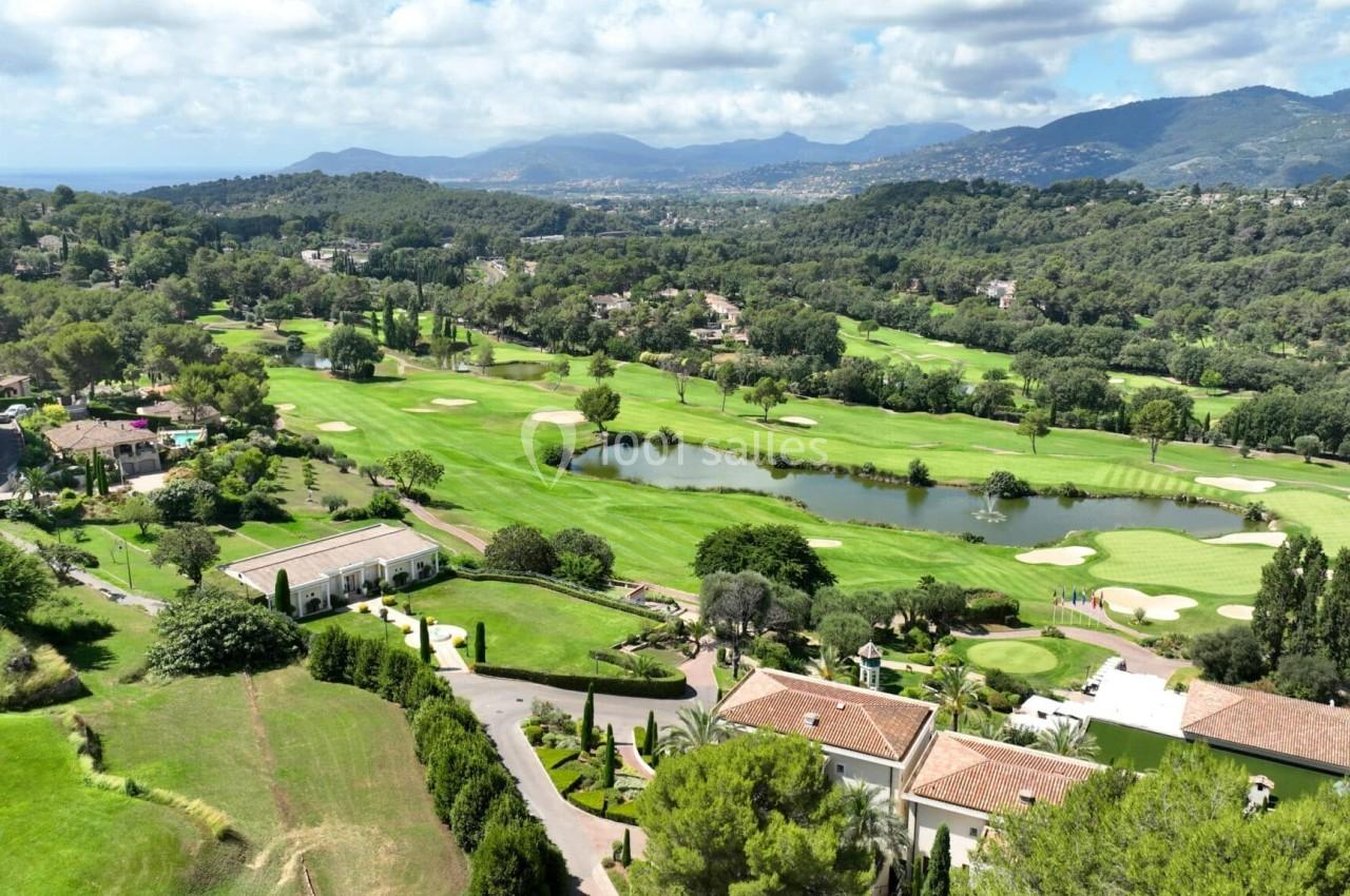 Vue aérienne d'un terrain de golf verdoyant avec un lac, entouré de collines et de bâtiments résidentiels.