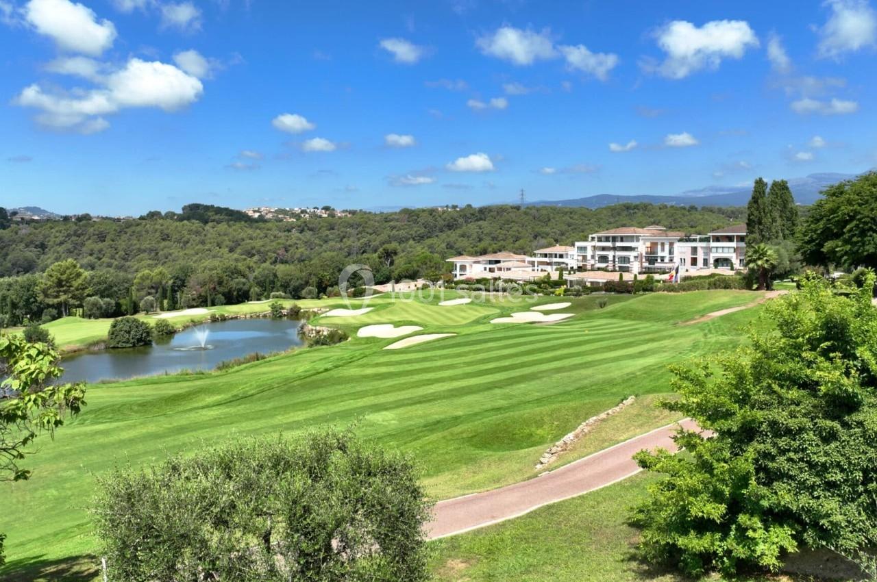 Vue d'un terrain de golf verdoyant avec un lac, des bunkers de sable, des bâtiments en arrière-plan et un ciel bleu.