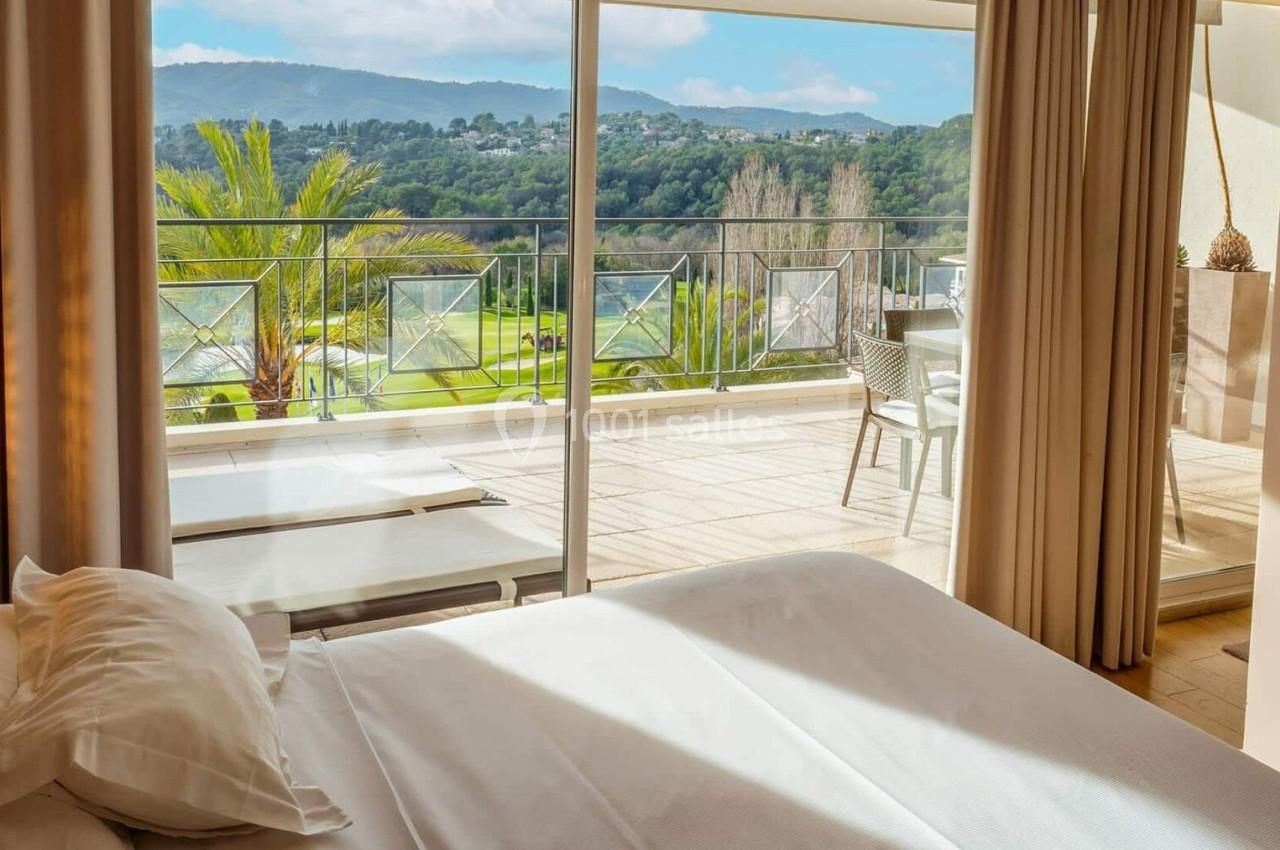 Chambre lumineuse avec lit double, rideaux ouverts sur une terrasse offrant une vue sur des collines verdoyantes.