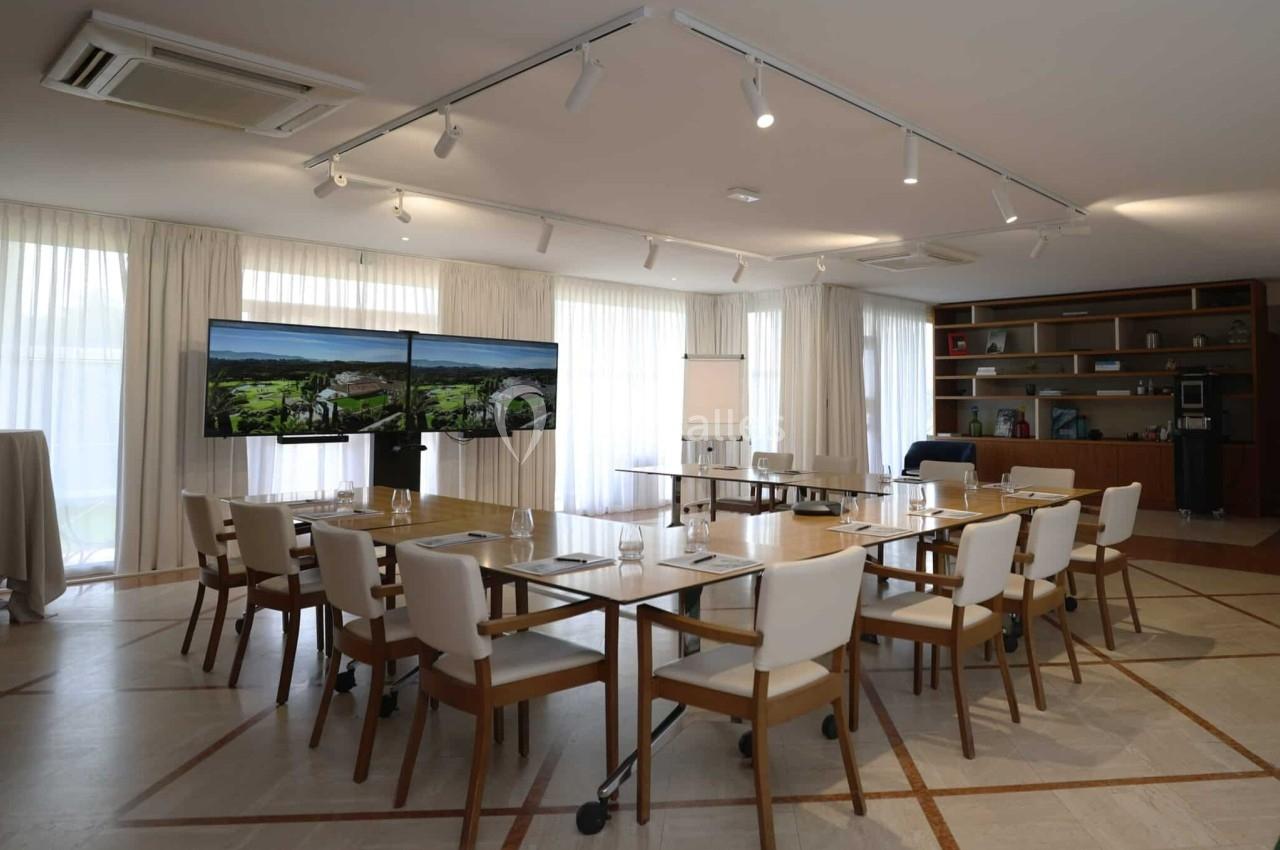Salle de réunion lumineuse avec tables en bois, chaises blanches, écrans, paperboard et étagères en arrière-plan.