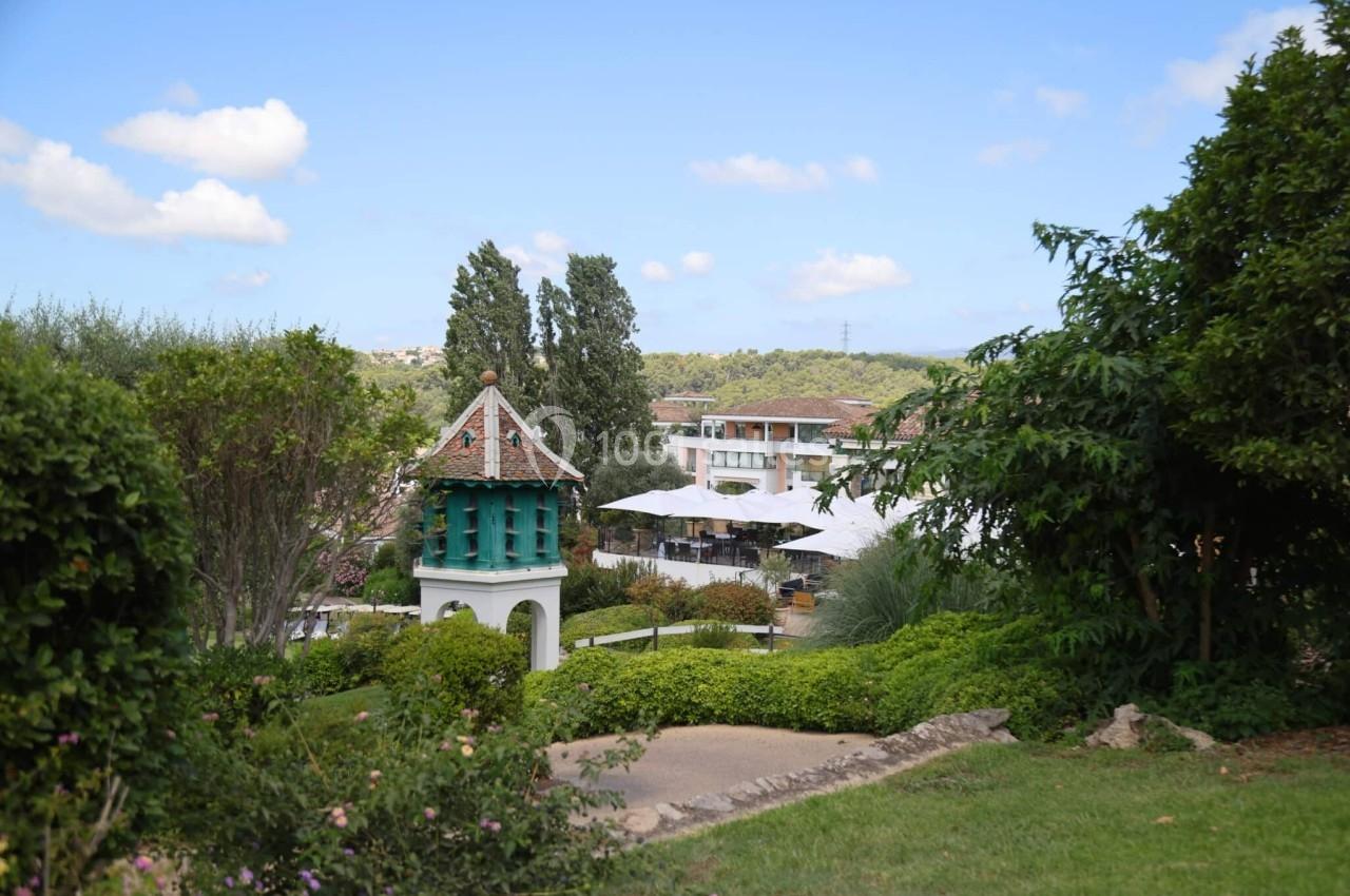 Jardin verdoyant avec un petit pigeonnier blanc et vert, des arbres et des bâtiments en arrière-plan sous un ciel bleu.