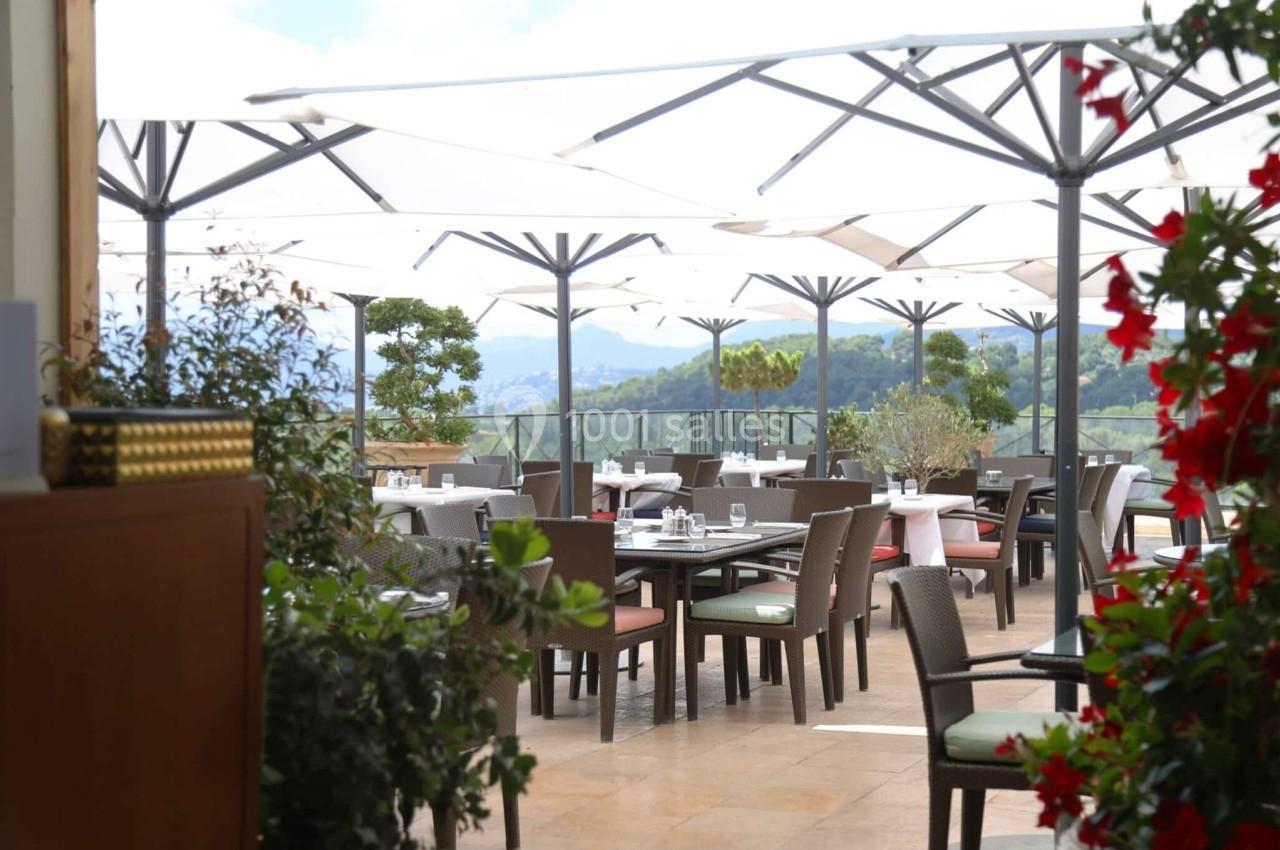 Terrasse d'un restaurant avec tables dressées, chaises en osier et parasols, entourée de plantes et vue sur des collines.
