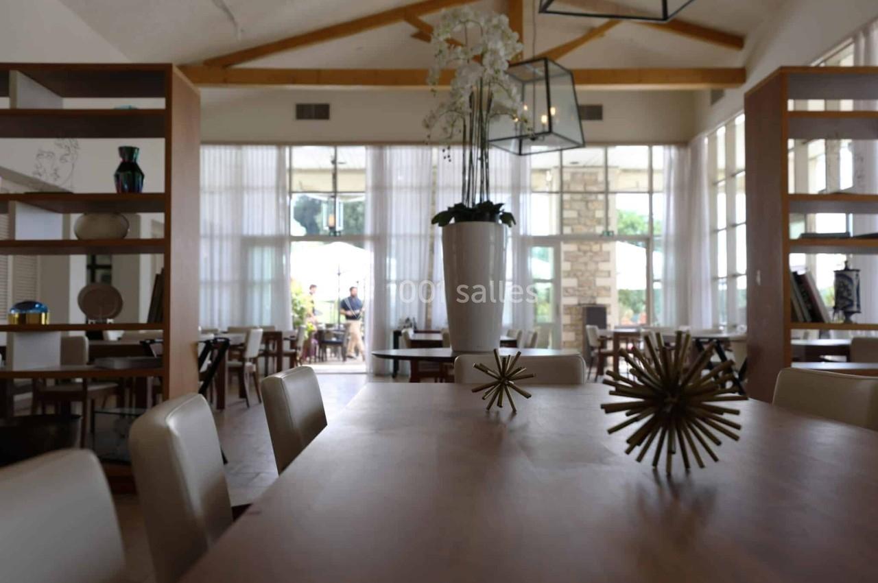 Salle de restaurant lumineuse avec grandes baies vitrées, tables en bois, chaises claires et décoration moderne.