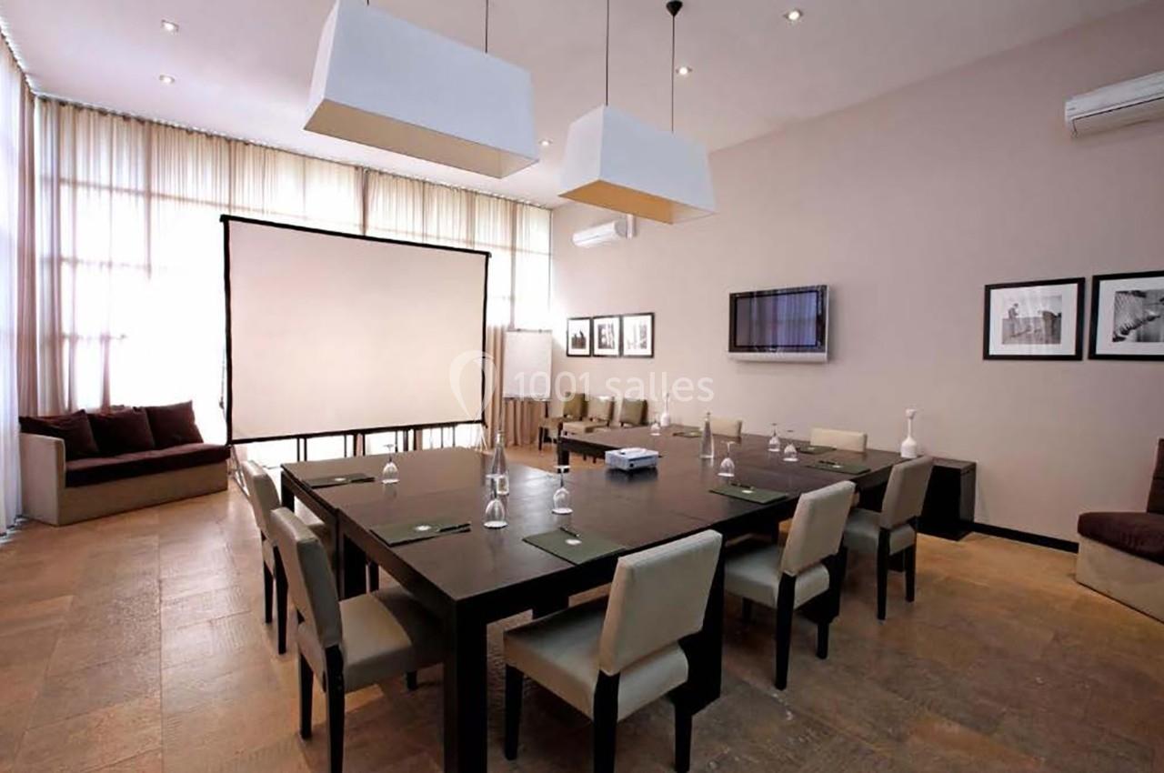 Salle de réunion lumineuse avec une grande table, des chaises, un écran de projection et des cadres accrochés au mur.