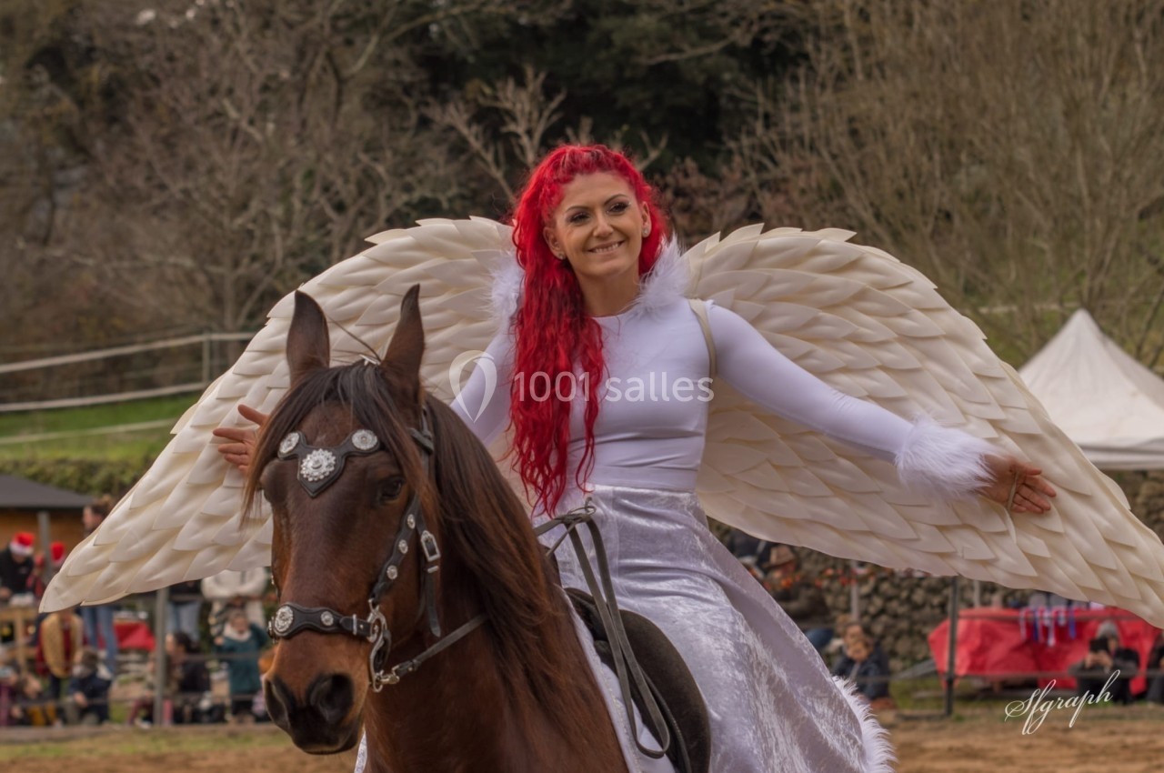 Femme aux cheveux rouges, vêtue d'ailes blanches, montant un cheval brun lors d'un événement en plein air.