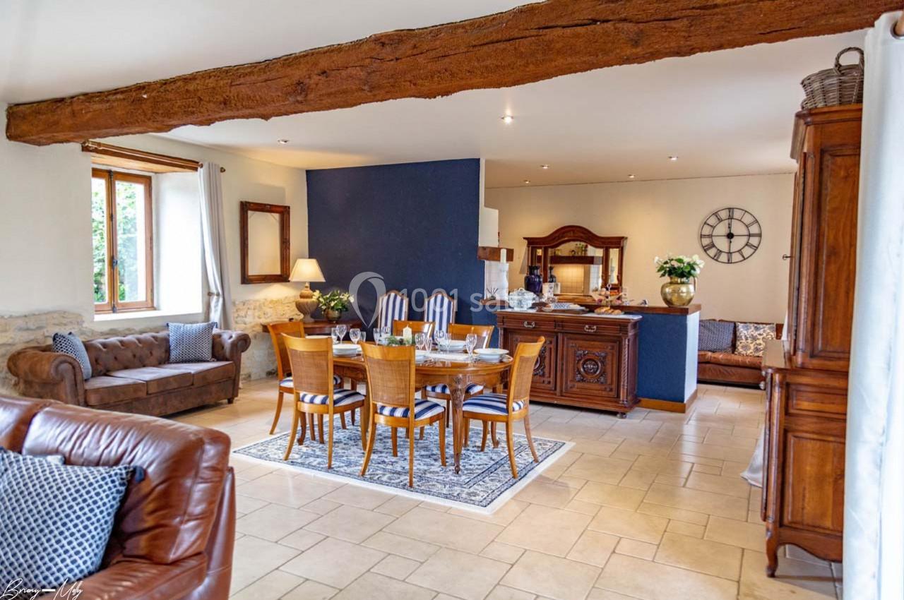 Salle à manger lumineuse avec table en bois, chaises rembourrées, poutre apparente et mobilier rustique.