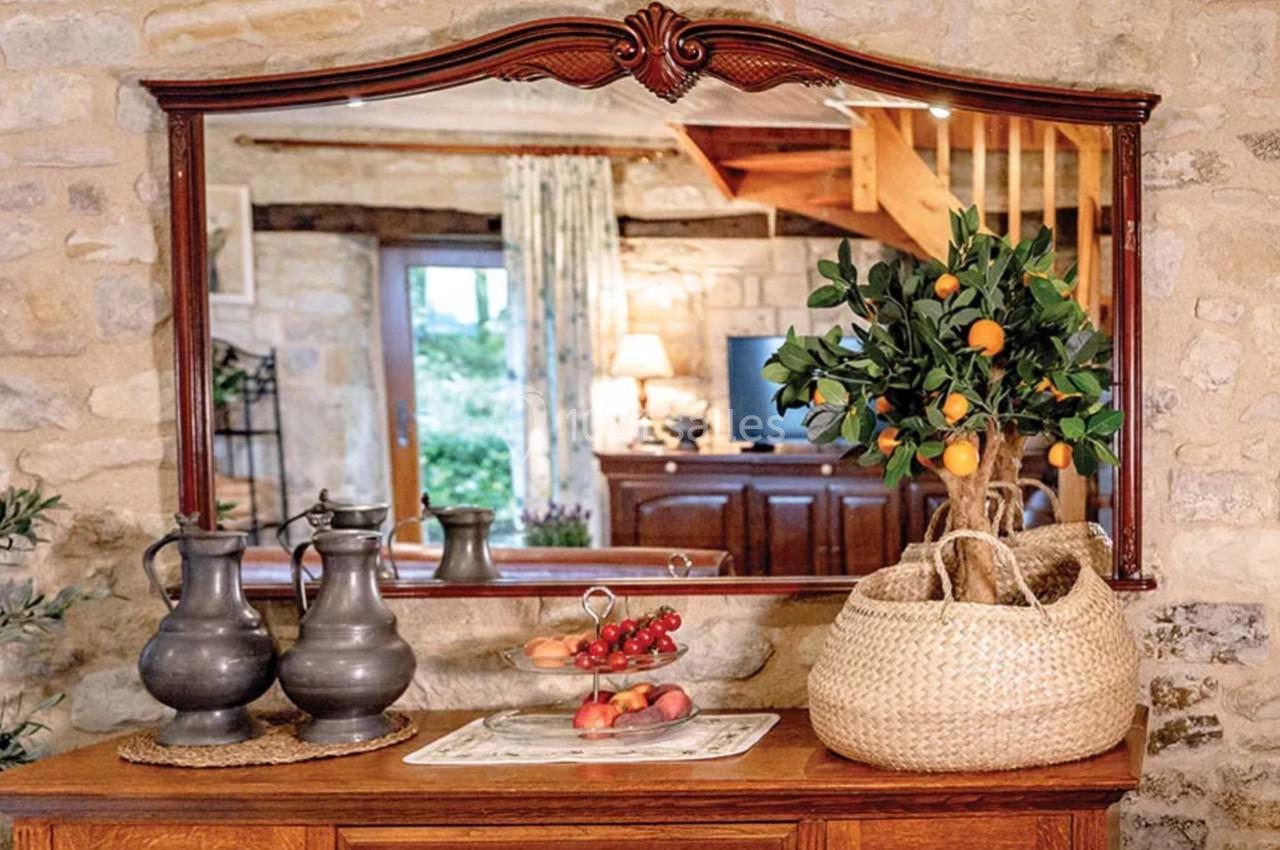 Un miroir encadré en bois reflète un intérieur rustique avec des murs en pierre, une table décorée de fruits et un panier.