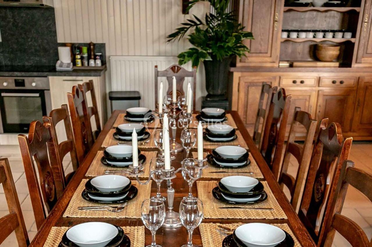Table en bois dressée avec vaisselle, verres et chandeliers, entourée de chaises dans une salle à manger rustique.
