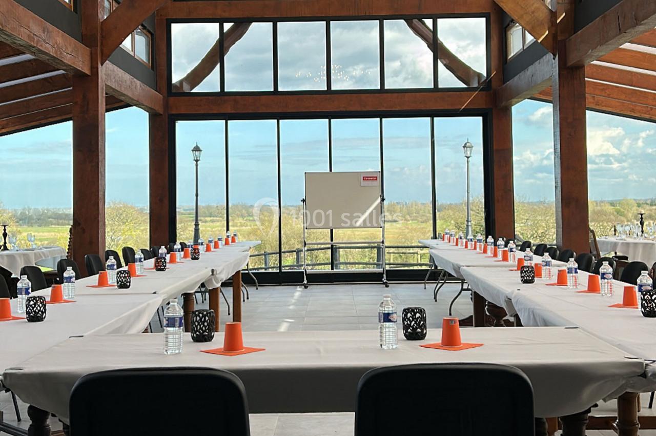 Salle de réunion lumineuse avec de grandes baies vitrées, tables disposées en U, et un tableau blanc au centre.