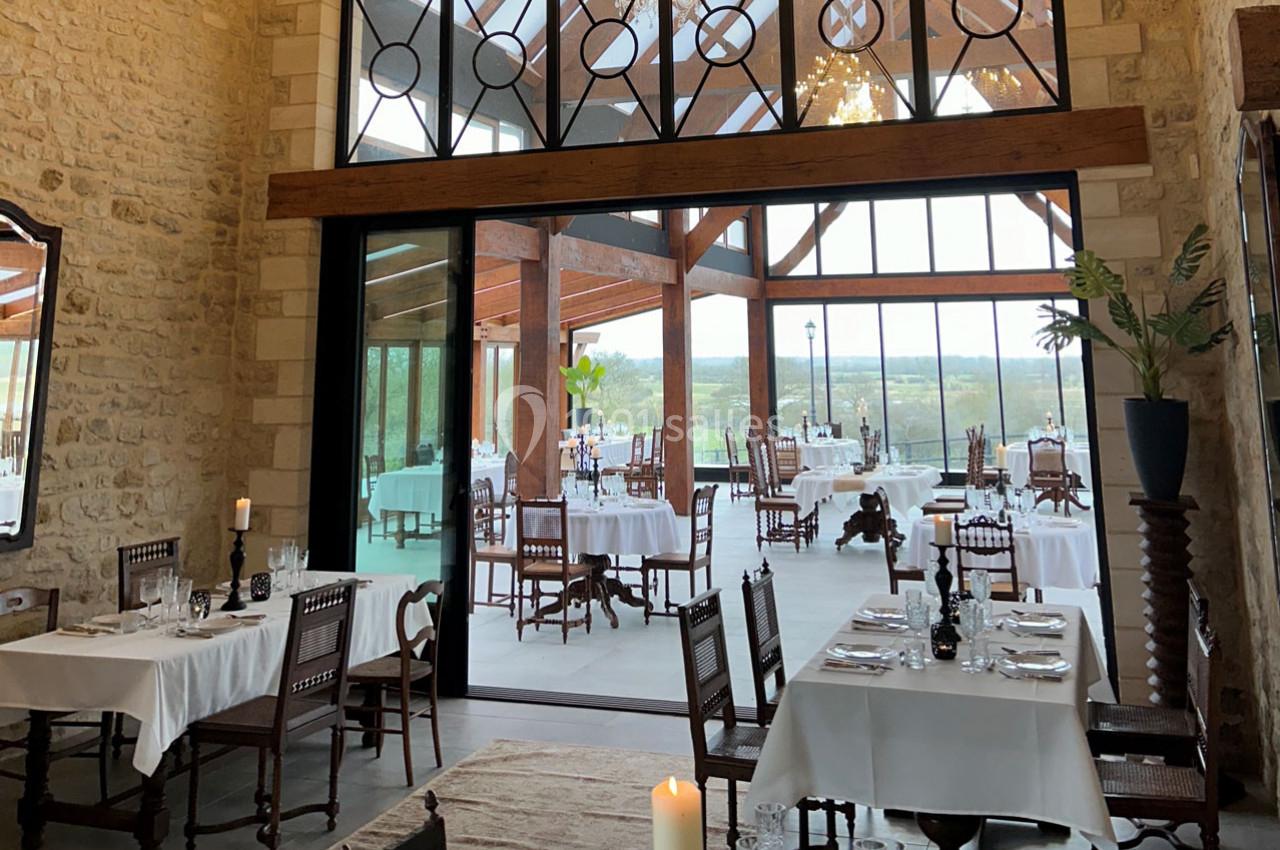 Salle de restaurant lumineuse avec murs en pierre, grandes baies vitrées et tables dressées avec nappes blanches.