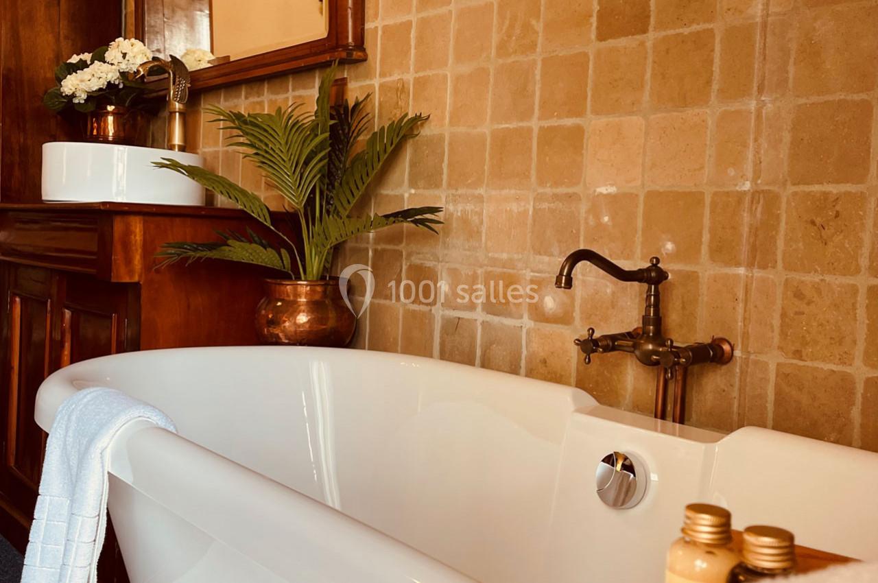 Baignoire blanche avec robinet en laiton, plantes décoratives et lavabo en arrière-plan sur un mur carrelé beige.