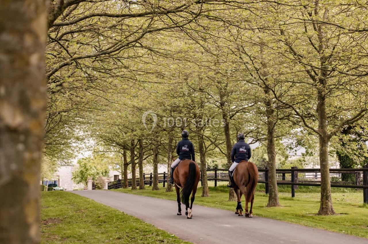 Deux cavaliers à cheval avancent sur une allée bordée d'arbres dans un cadre verdoyant.