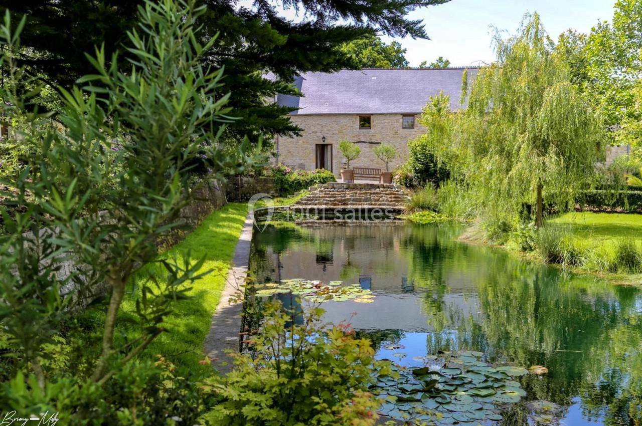 Étang bordé de végétation avec une maison en pierre et un jardin arboré en arrière-plan.