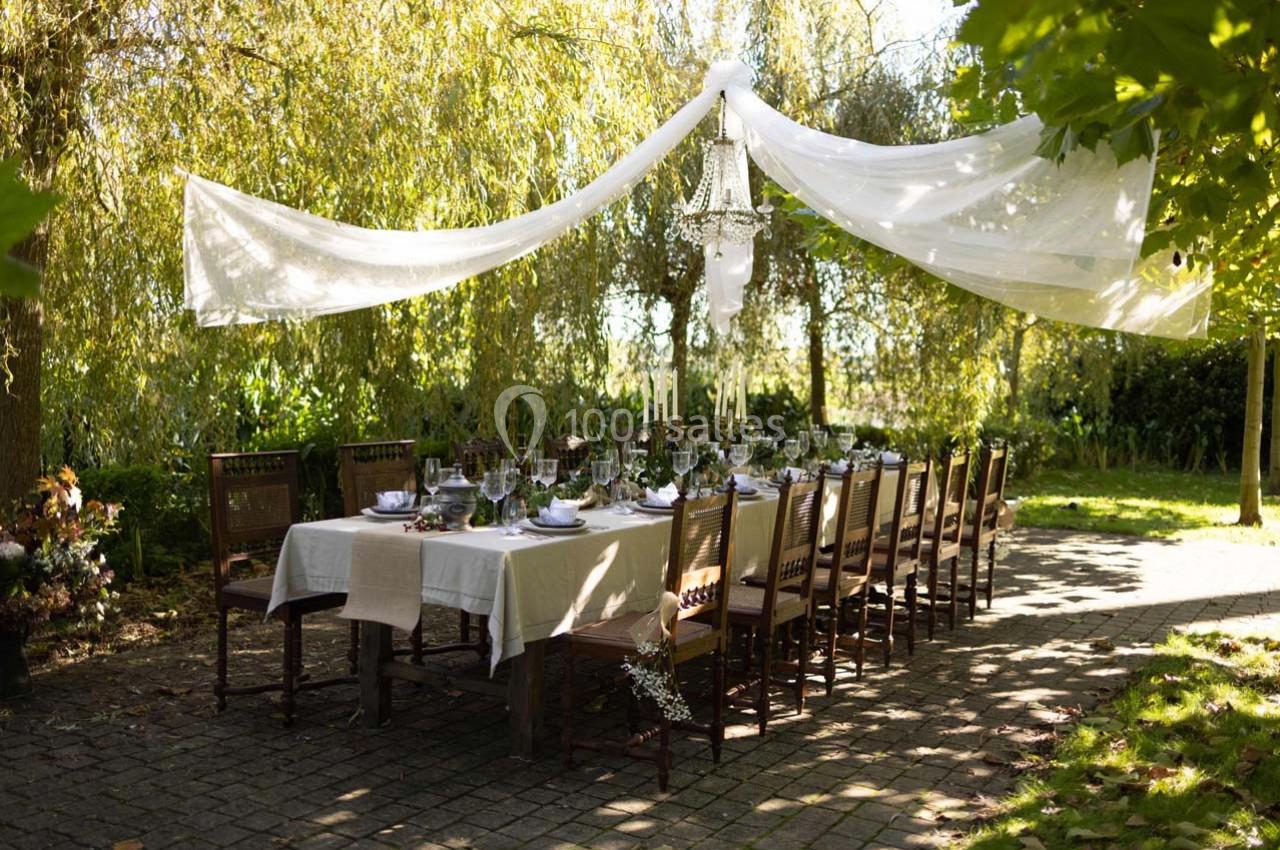Table dressée en extérieur sous un auvent de tissu blanc, entourée de chaises en bois et située dans un jardin ombragé.
