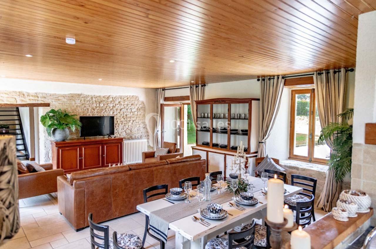 Salle à manger et salon lumineux avec mobilier en bois, canapé en cuir, murs en pierre et grandes fenêtres.