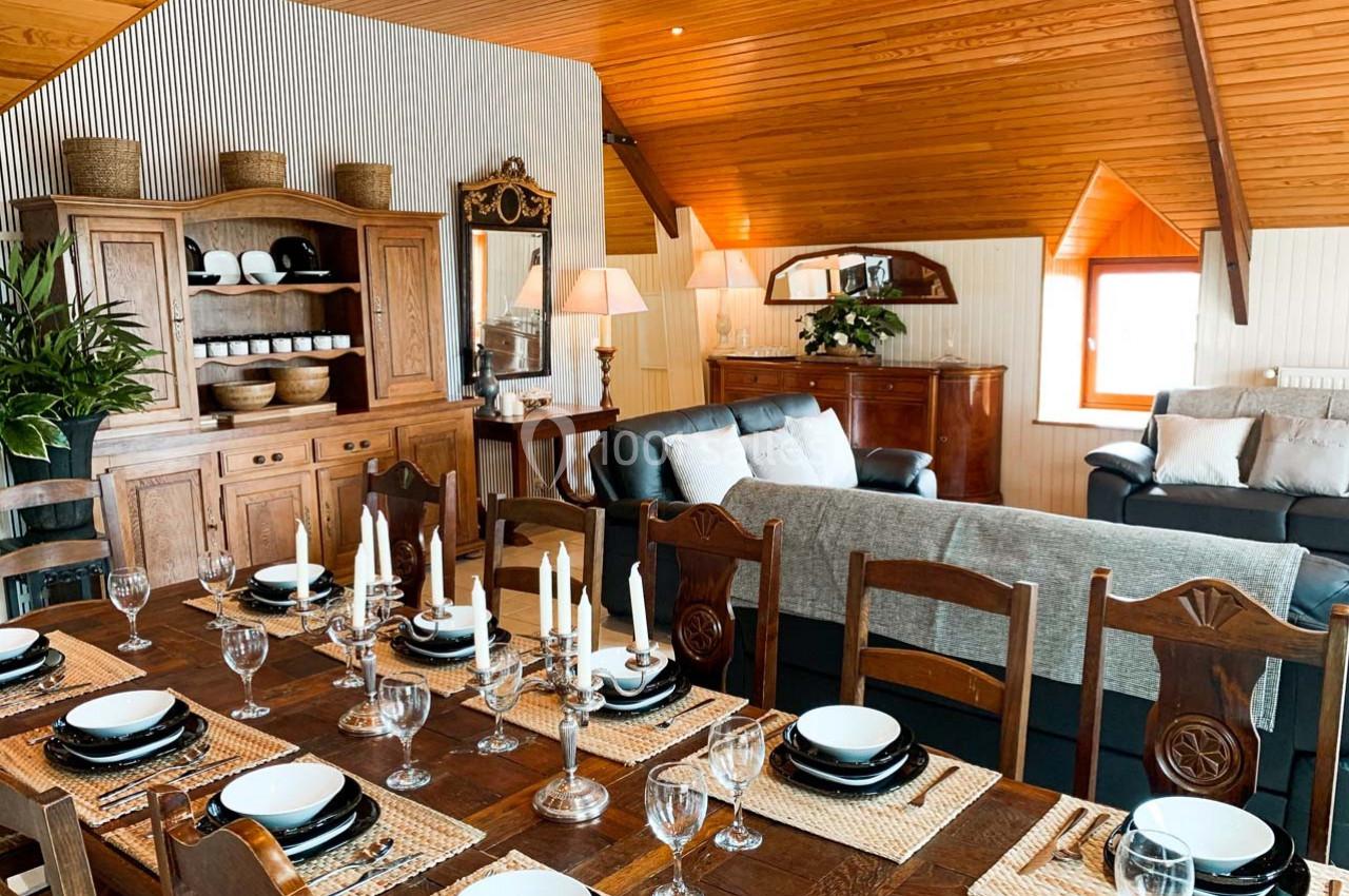 Salle à manger rustique avec table en bois dressée, chaises assorties, et salon confortable en arrière-plan.