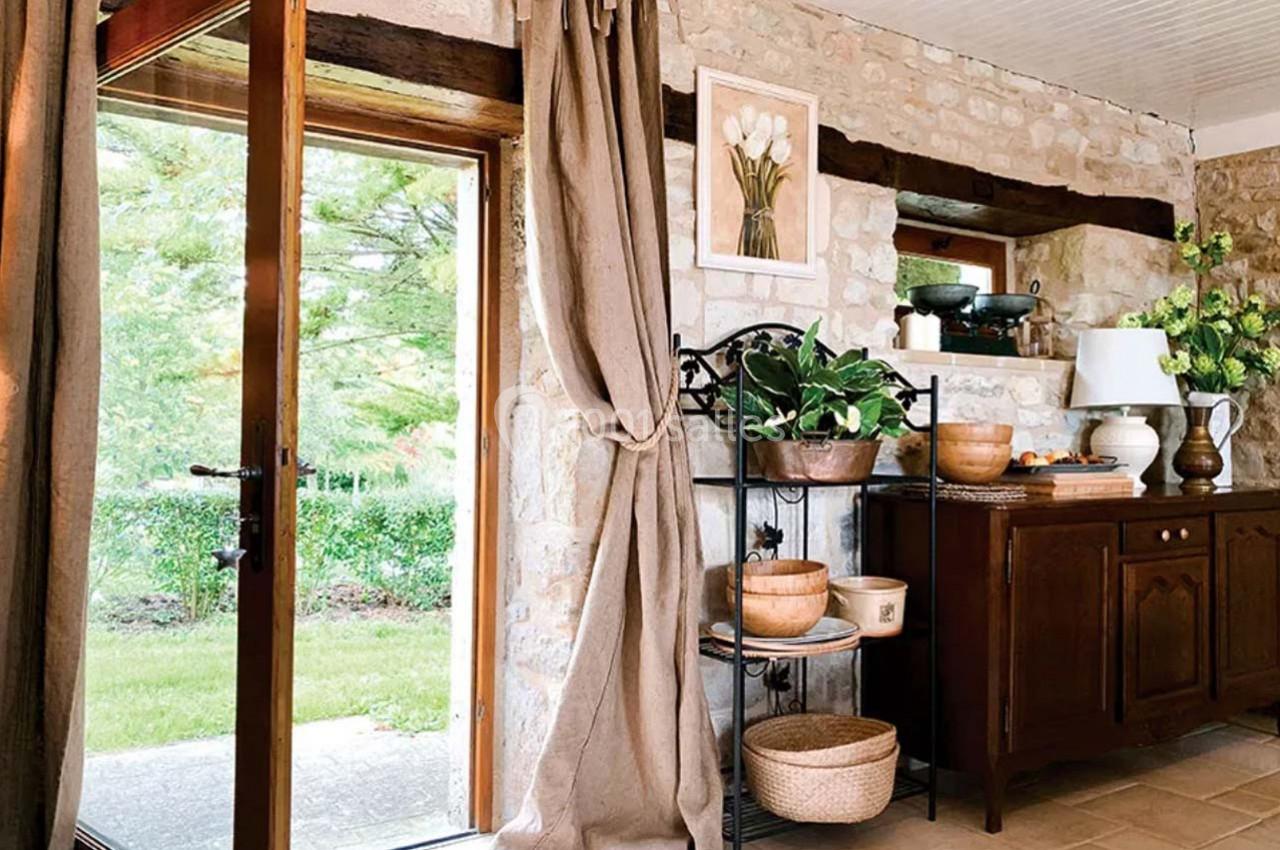 Intérieur rustique avec murs en pierre, meubles en bois, plantes et vue sur un jardin par une porte vitrée.