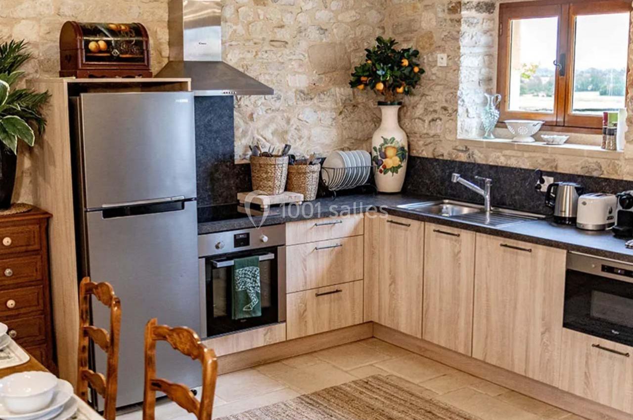 Cuisine rustique avec meubles en bois clair, électroménagers modernes, évier, et décoration en pierre apparente.