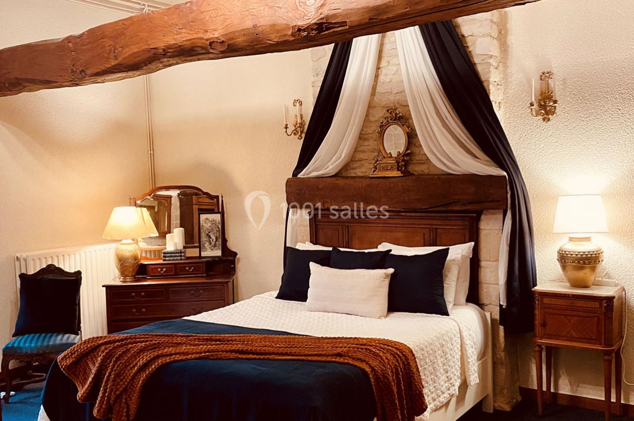 Chambre chaleureuse avec lit double, poutre apparente, draperies décoratives, meubles en bois et éclairage doux.