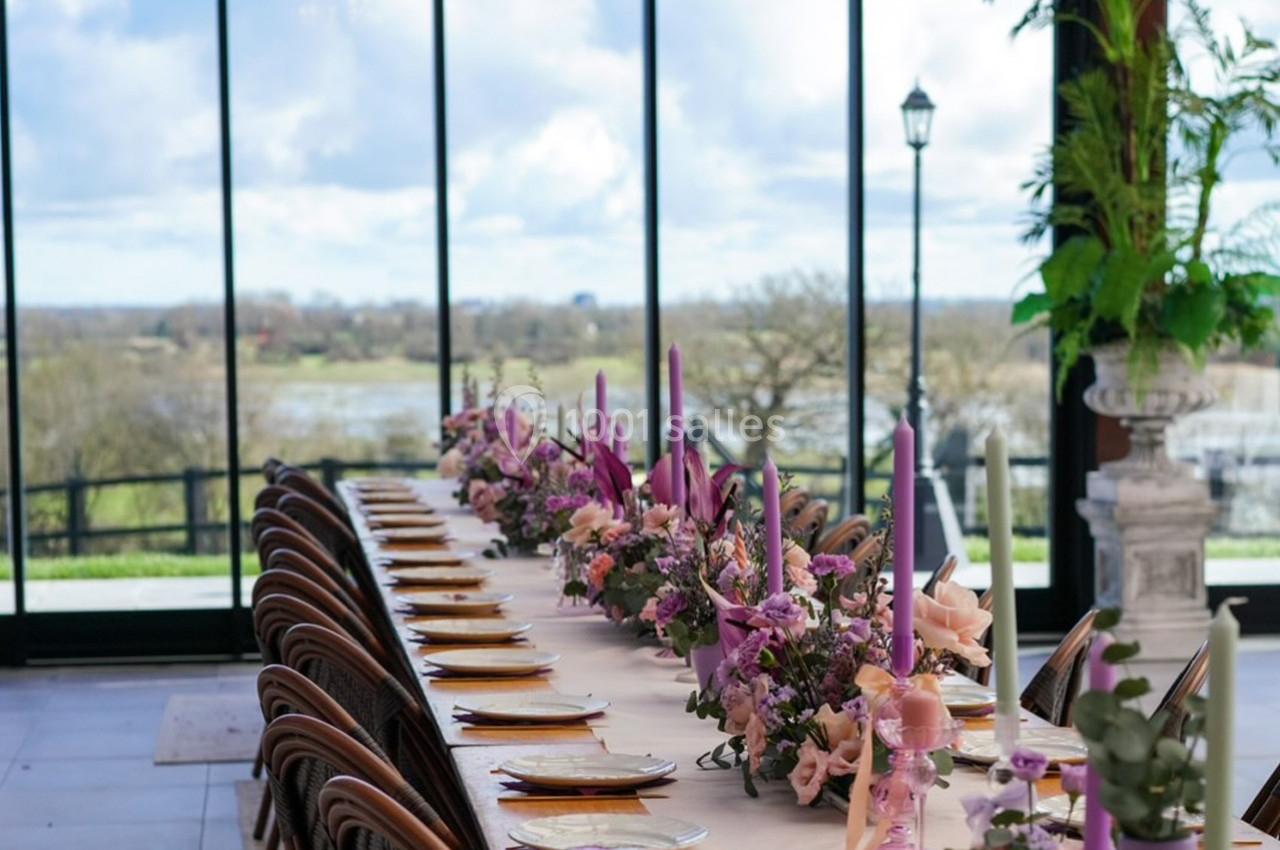 Table décorée avec des fleurs roses et des bougies colorées, placée devant de grandes baies vitrées avec vue sur un paysage.