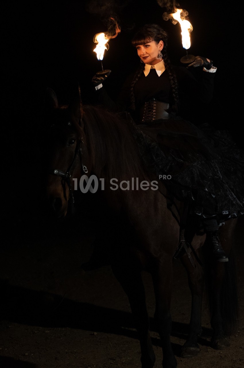 Femme en tenue sombre montée sur un cheval, tenant des torches enflammées dans un environnement nocturne.