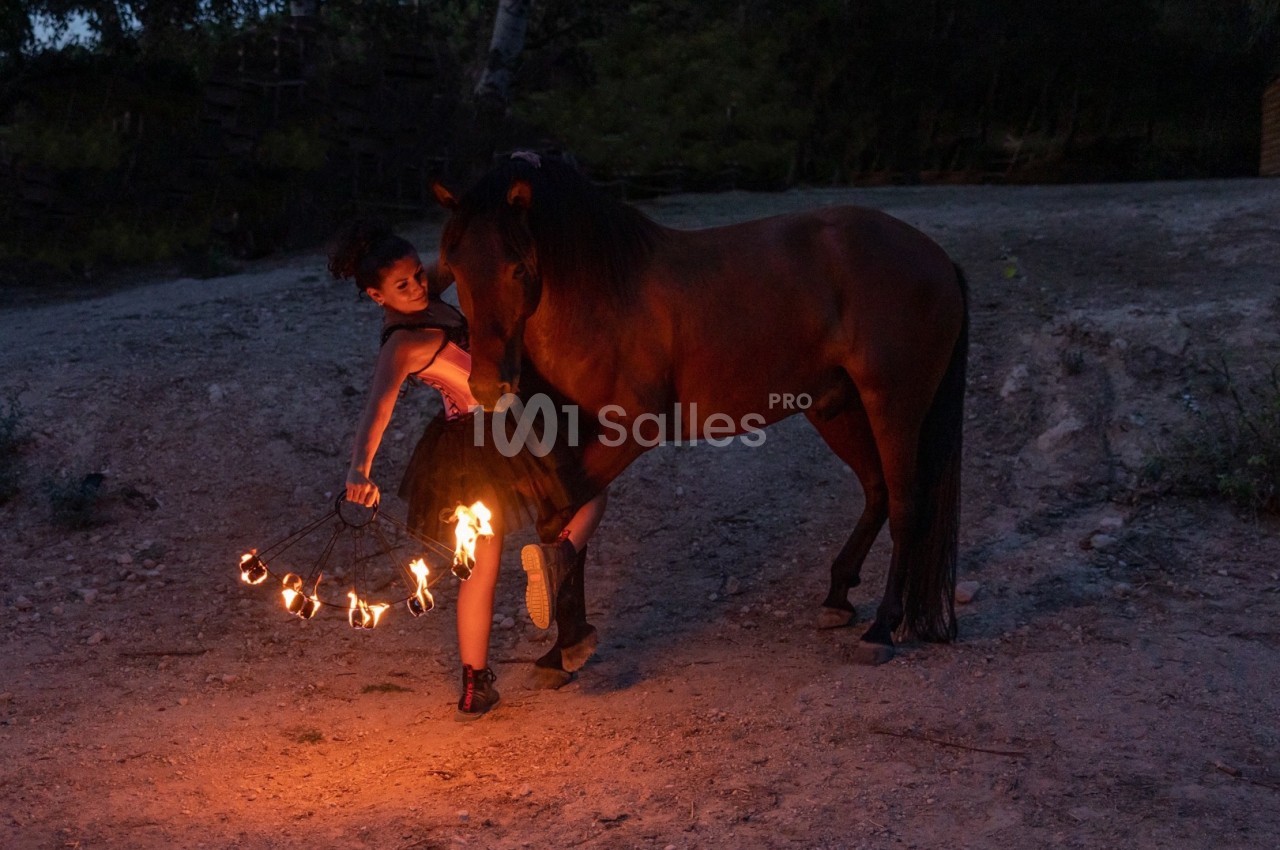 Une femme jongle avec des torches enflammées près d'un cheval dans un environnement extérieur sombre.
