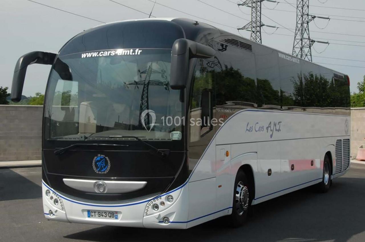 Autocar blanc stationné sur un parking, avec des inscriptions sur la carrosserie et des pylônes électriques en arrière-plan.