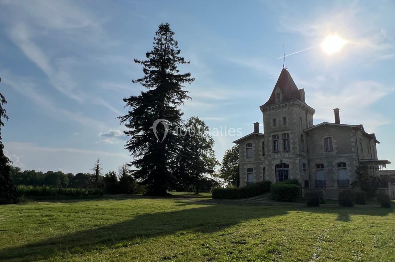 Manoir en pierre avec une tour, entouré d'un grand jardin verdoyant et d'arbres, sous un ciel ensoleillé.