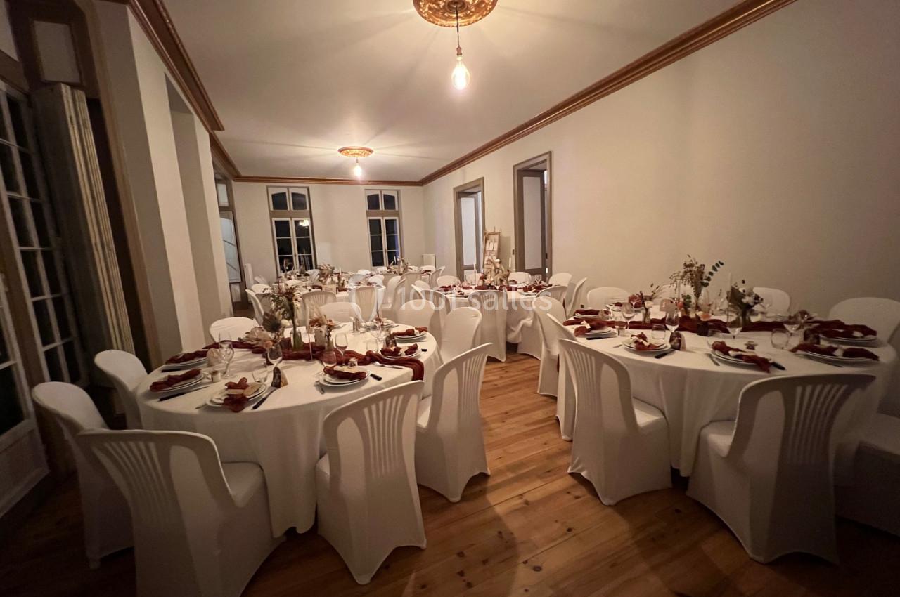 Salle de réception avec tables rondes dressées, nappes blanches, chaises en plastique et décorations florales.