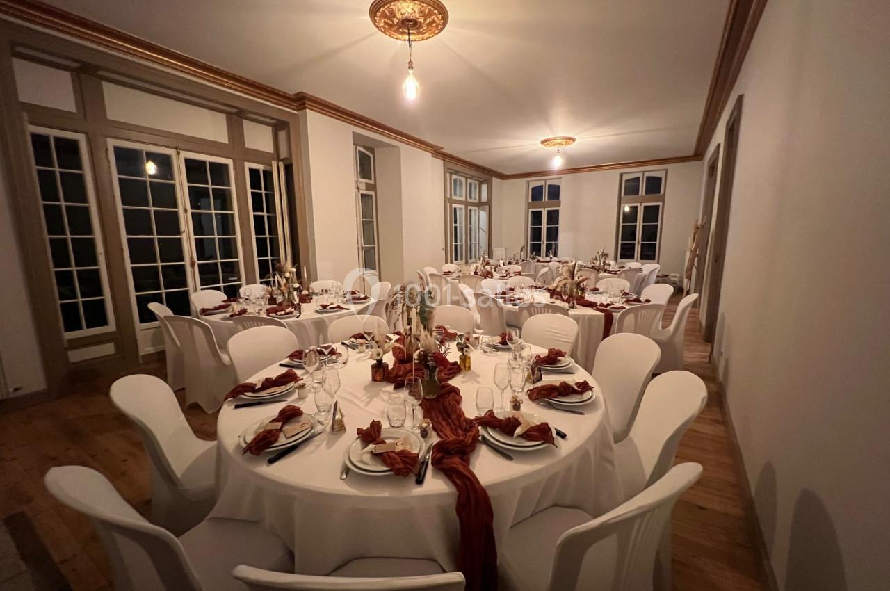Salle élégante avec tables rondes dressées, nappes blanches, serviettes rouges et décorations florales.