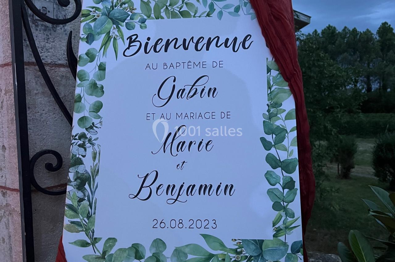 Panneau décoratif avec feuillage vert annonçant un baptême et un mariage, posé sur un chevalet en extérieur.