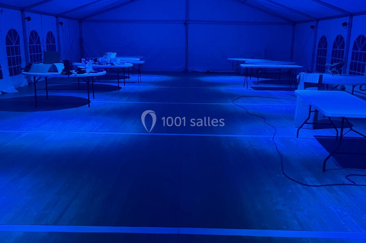 Intérieur d'une grande tente éclairée en bleu, avec des tables vides alignées sur un sol en bois.