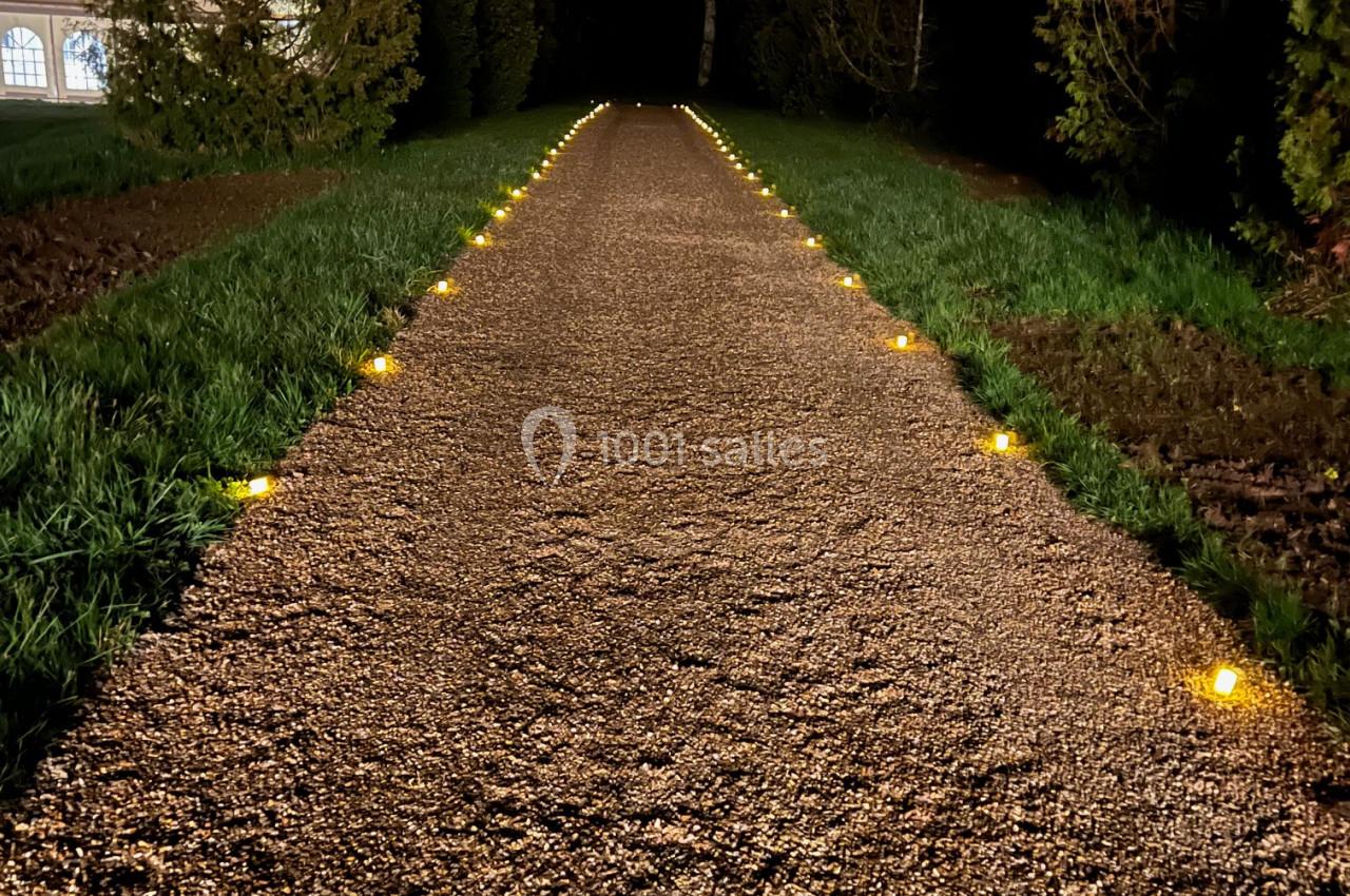 Chemin de gravier éclairé par des lumières au sol, bordé d'arbres et d'herbe, dans un environnement nocturne.