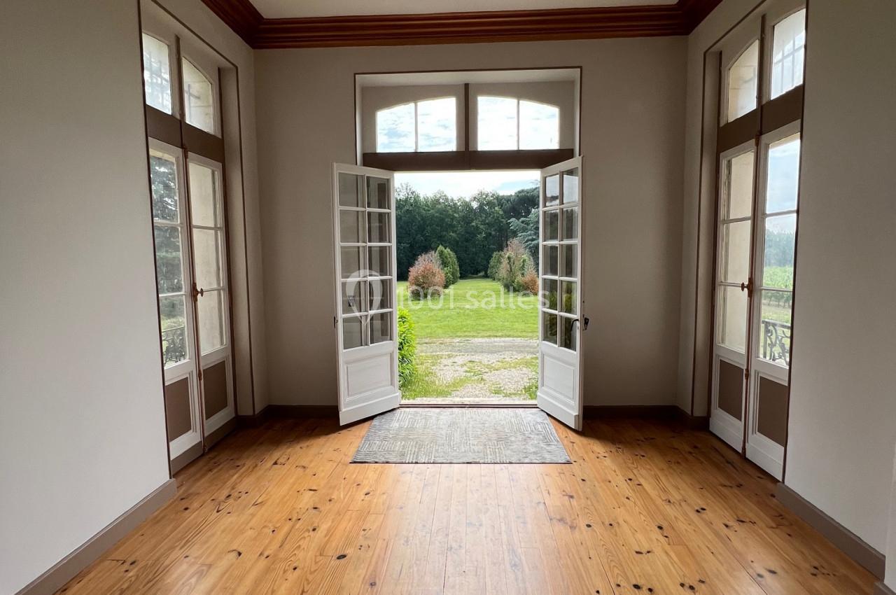 Salle lumineuse avec parquet en bois, portes-fenêtres ouvertes donnant sur un jardin verdoyant et un ciel partiellement…