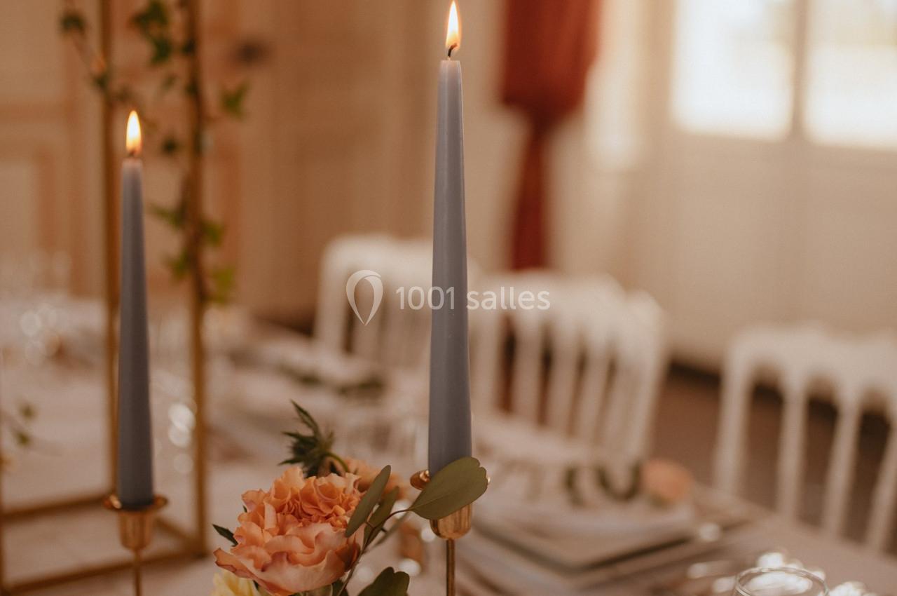 Bougies allumées et fleurs dans un vase décorent une table élégamment dressée dans une pièce lumineuse.