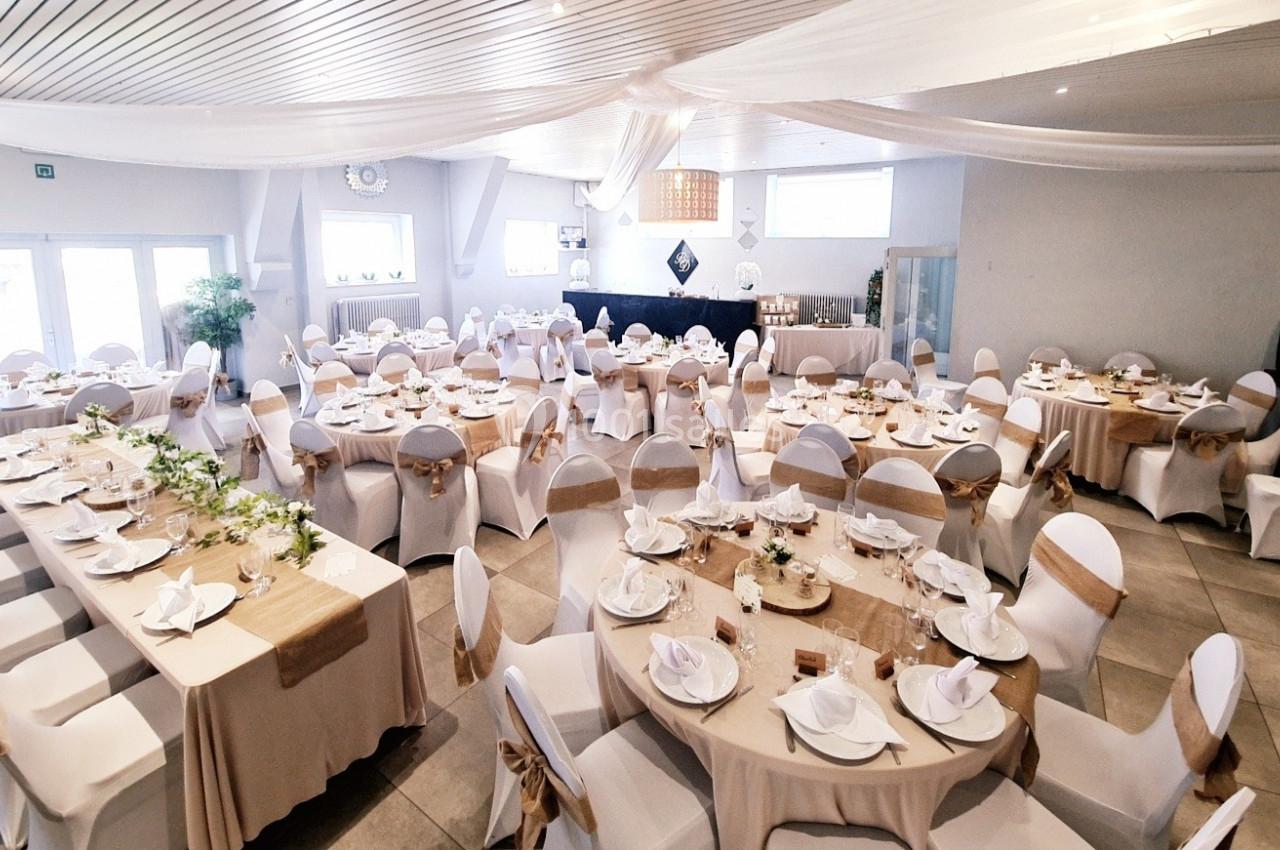 Salle de réception décorée avec des tables dressées, nappes beiges, chaises blanches et rubans dorés.