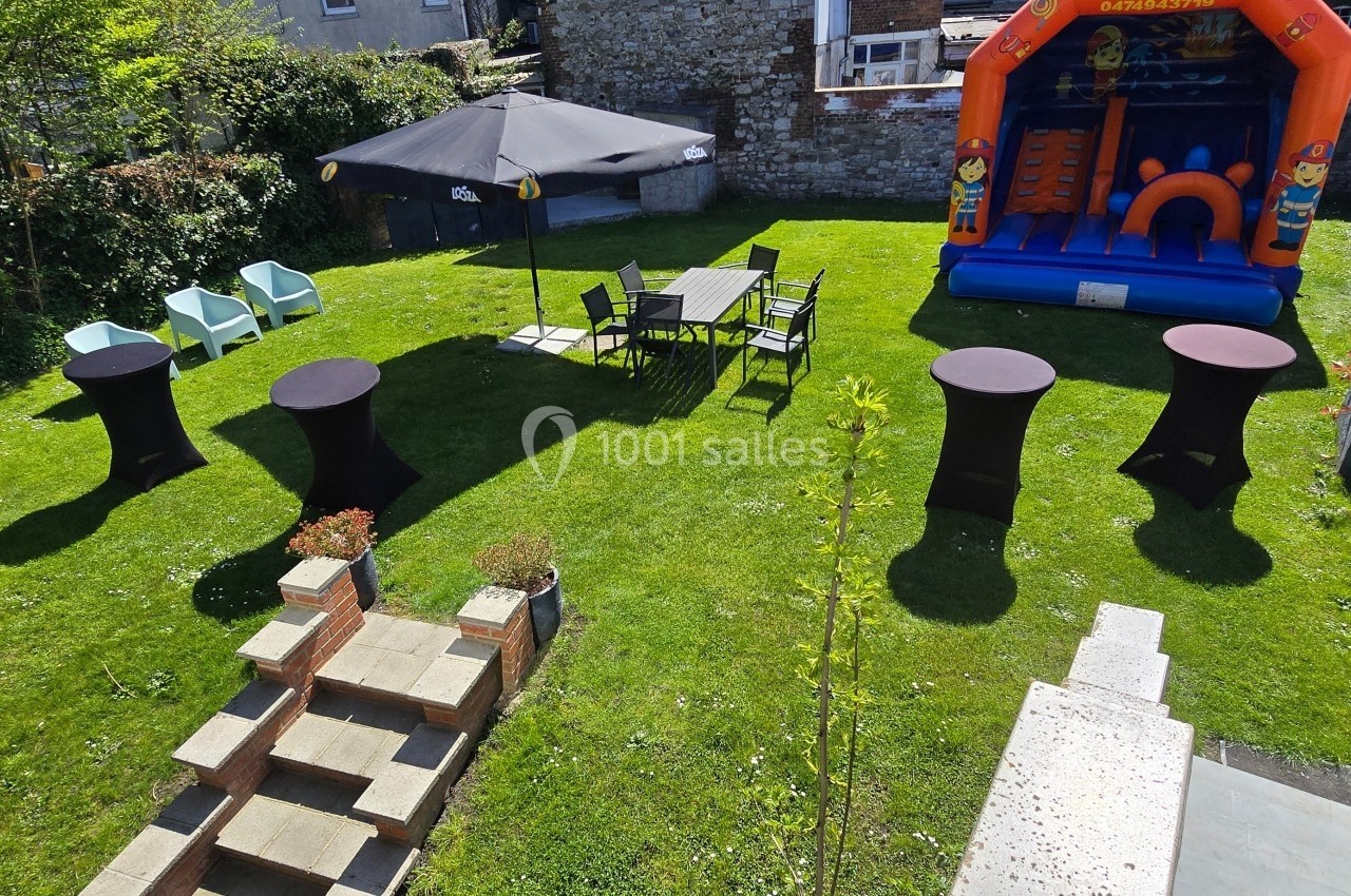 Jardin aménagé avec tables, chaises, parasol, structures gonflables et espaces verts entourés de murs et végétation.