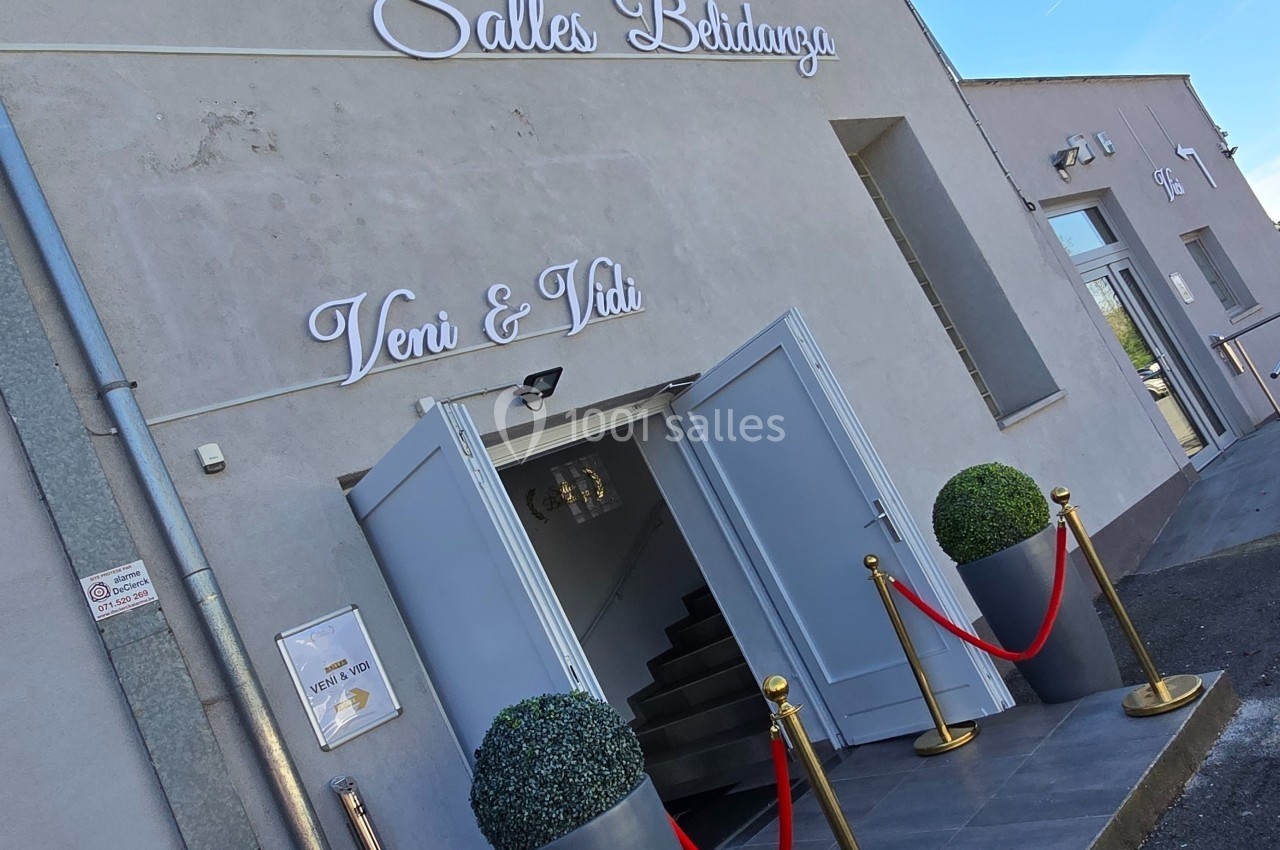 Façade d'une salle de réception avec portes ouvertes, tapis rouge, poteaux de guidage et deux plantes décoratives.