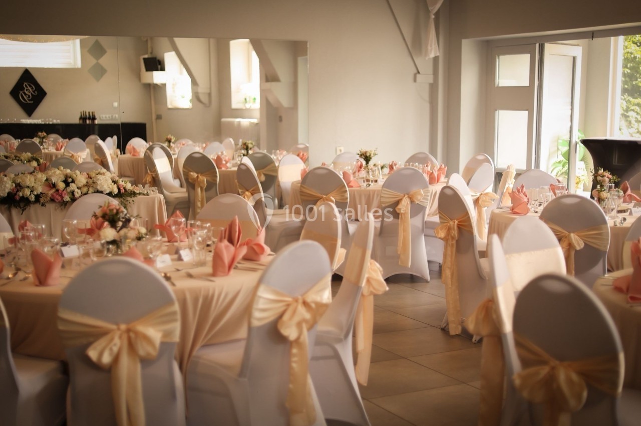 Salle de réception décorée avec des tables rondes, nappes beige, chaises ornées de nœuds et compositions florales.