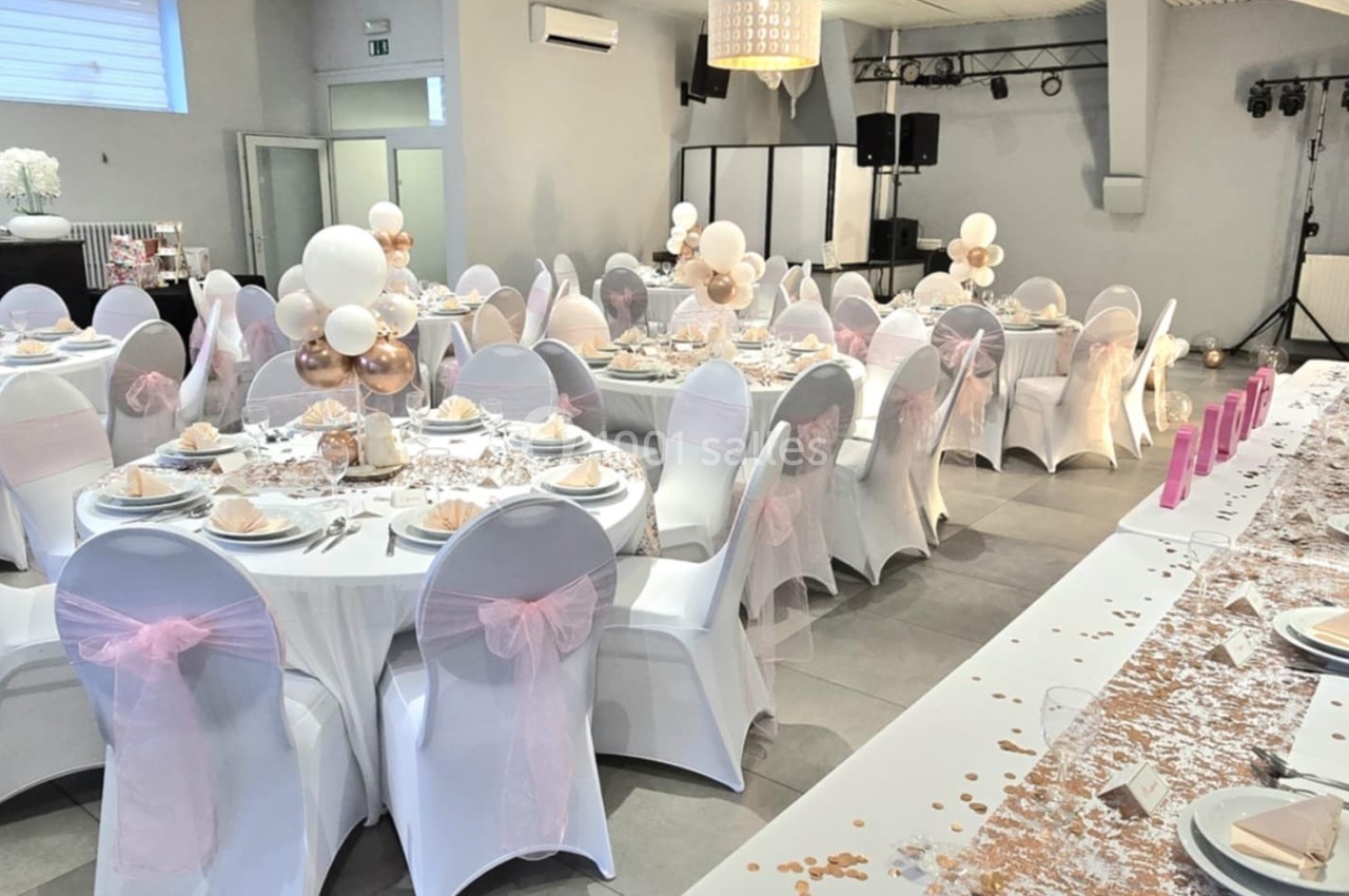 Salle de réception décorée pour un événement, avec tables rondes dressées, nappes blanches et décorations roses et dorées.