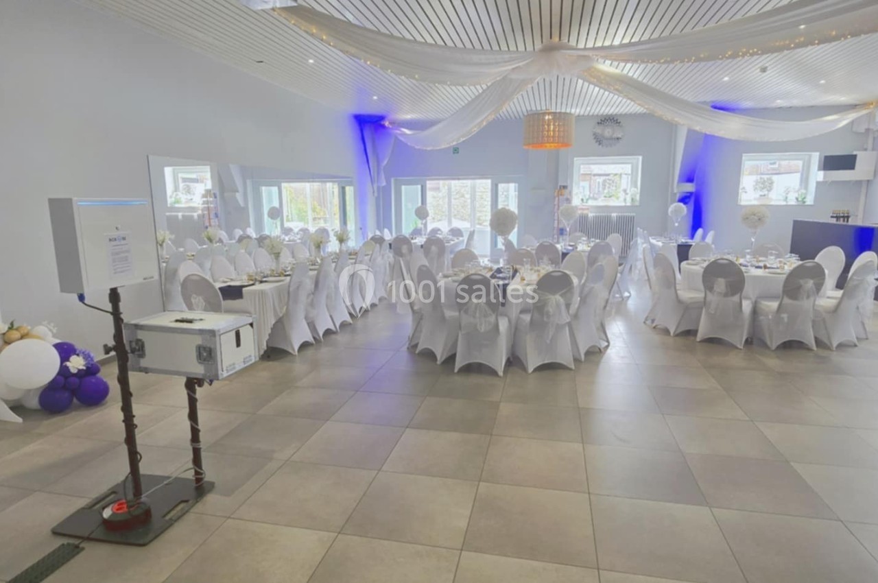Salle de réception décorée avec des tables rondes dressées, des chaises blanches et des ballons en arrière-plan.