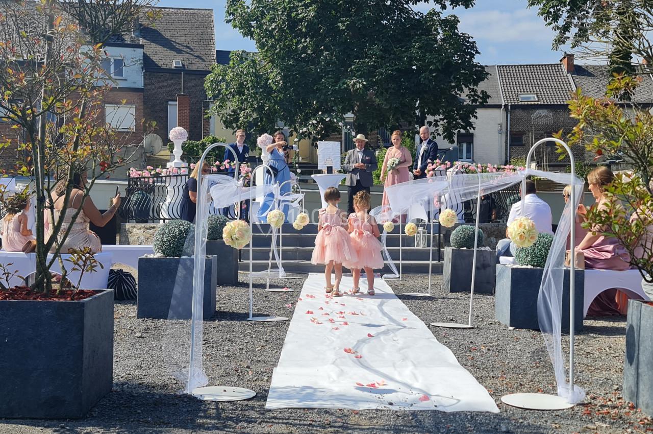 Deux enfants en robes roses marchent sur une allée blanche décorée de fleurs lors d'une cérémonie en extérieur.